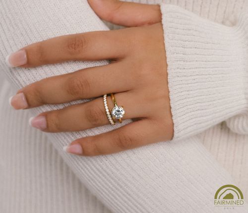 Hand model wears Round Diamond Solitaire Engagement Ring Mounting in Fairmined Yellow Gold with half-pave wedding band, from RIVA Precision