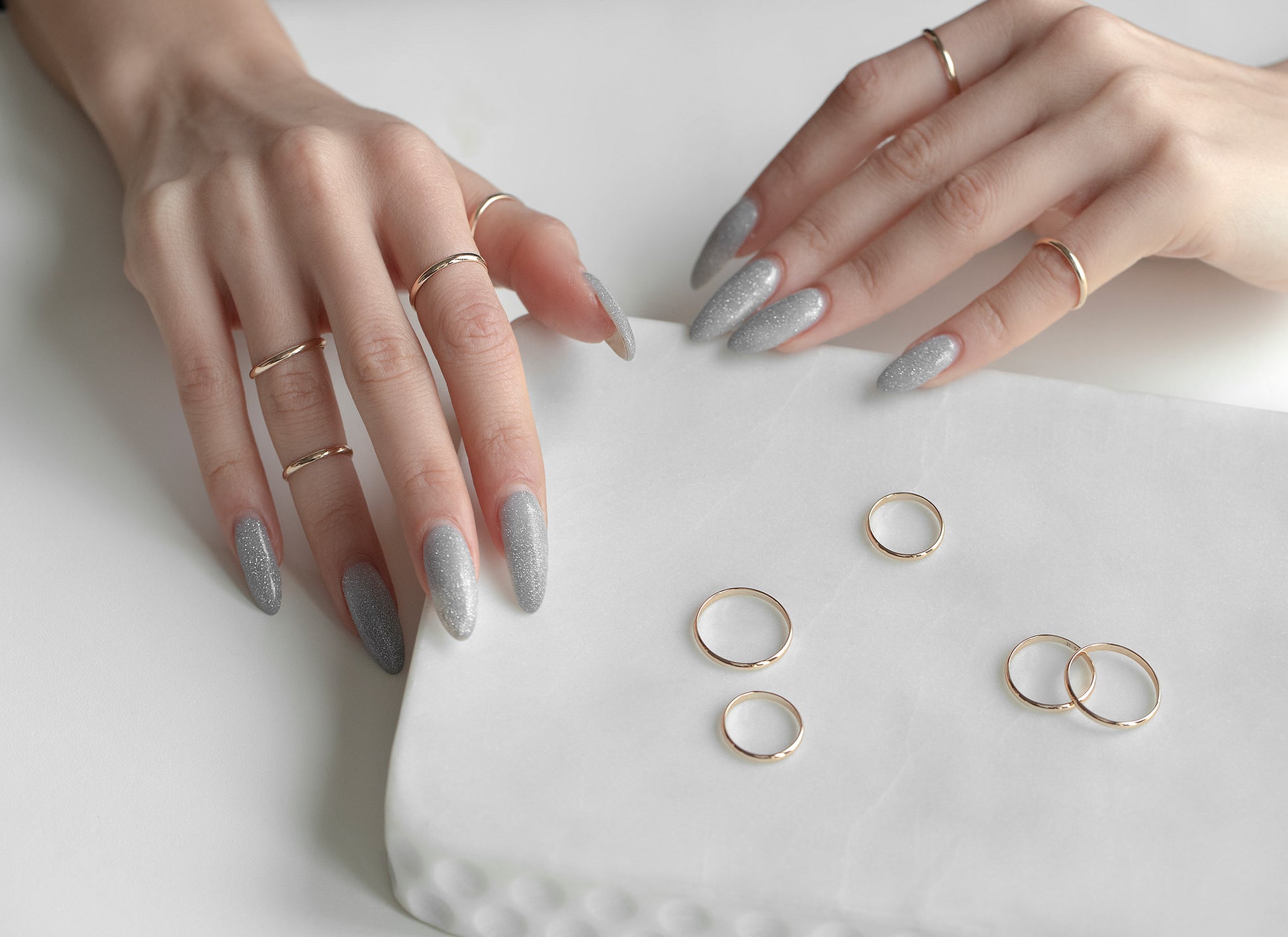 Hands with gray nail polish holding a white cushion with gold rings on a light background