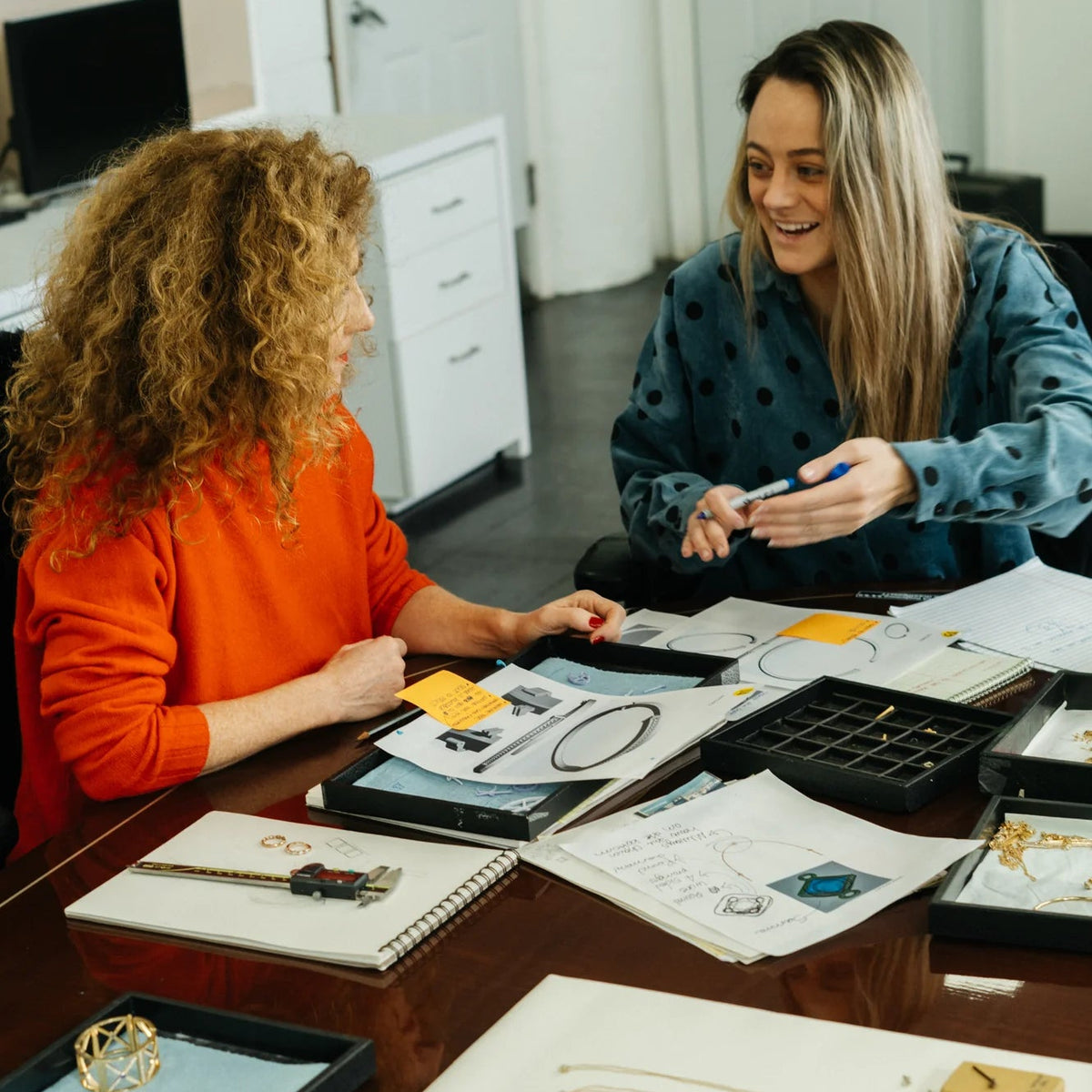 Two women in an office setting discussing jewelry on a table.