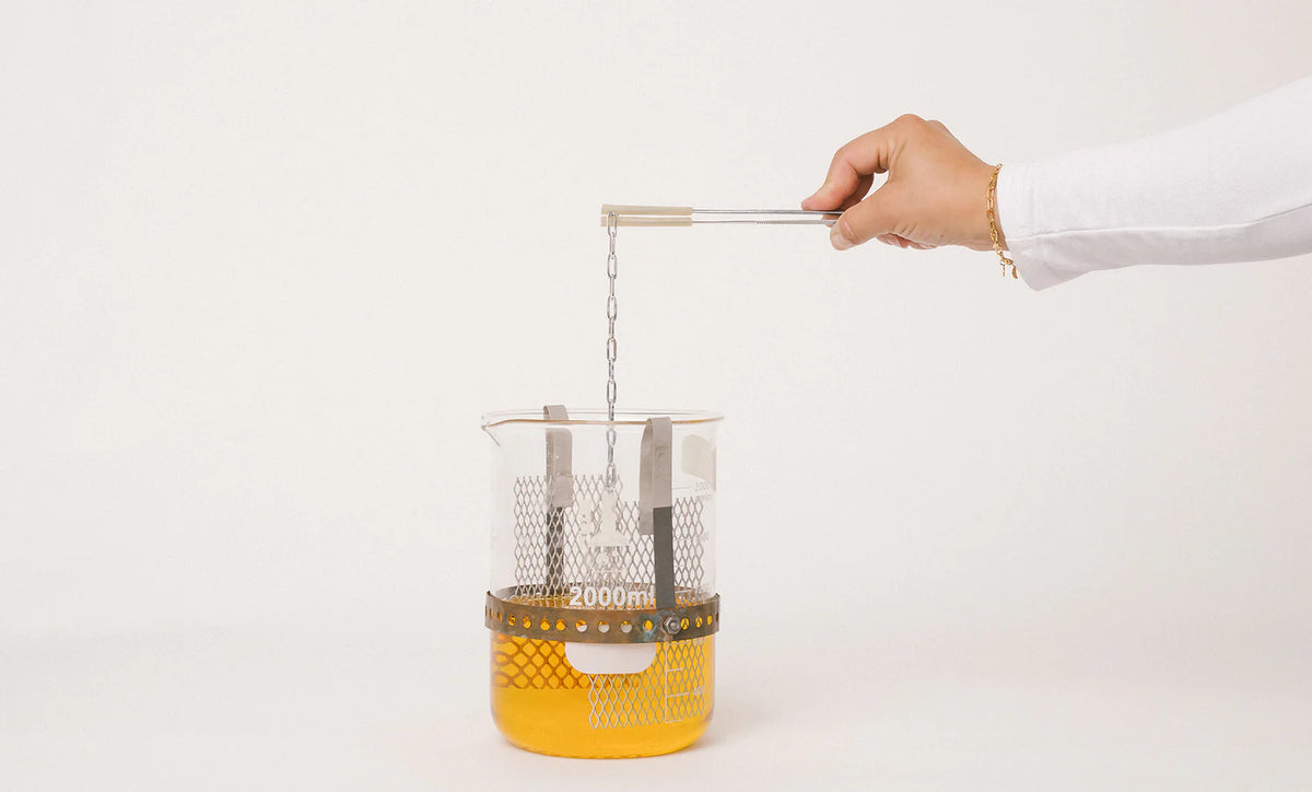 Person plating metal strainer over a container with yellow liquid on a white background