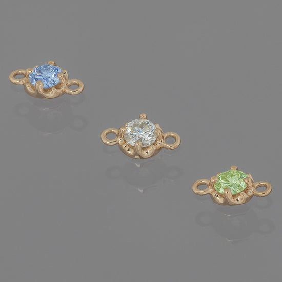 Three prong-set birthstone connectors in gold, each with a different color gemstone, displayed against a grey background.