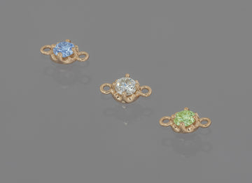 Three prong-set birthstone connectors in gold, each with a different color gemstone, displayed against a grey background.