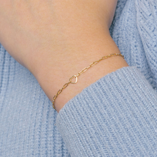 A gold heart-shaped wire charm connector worn as a bracelet on a person's wrist, with a sweater in the background.