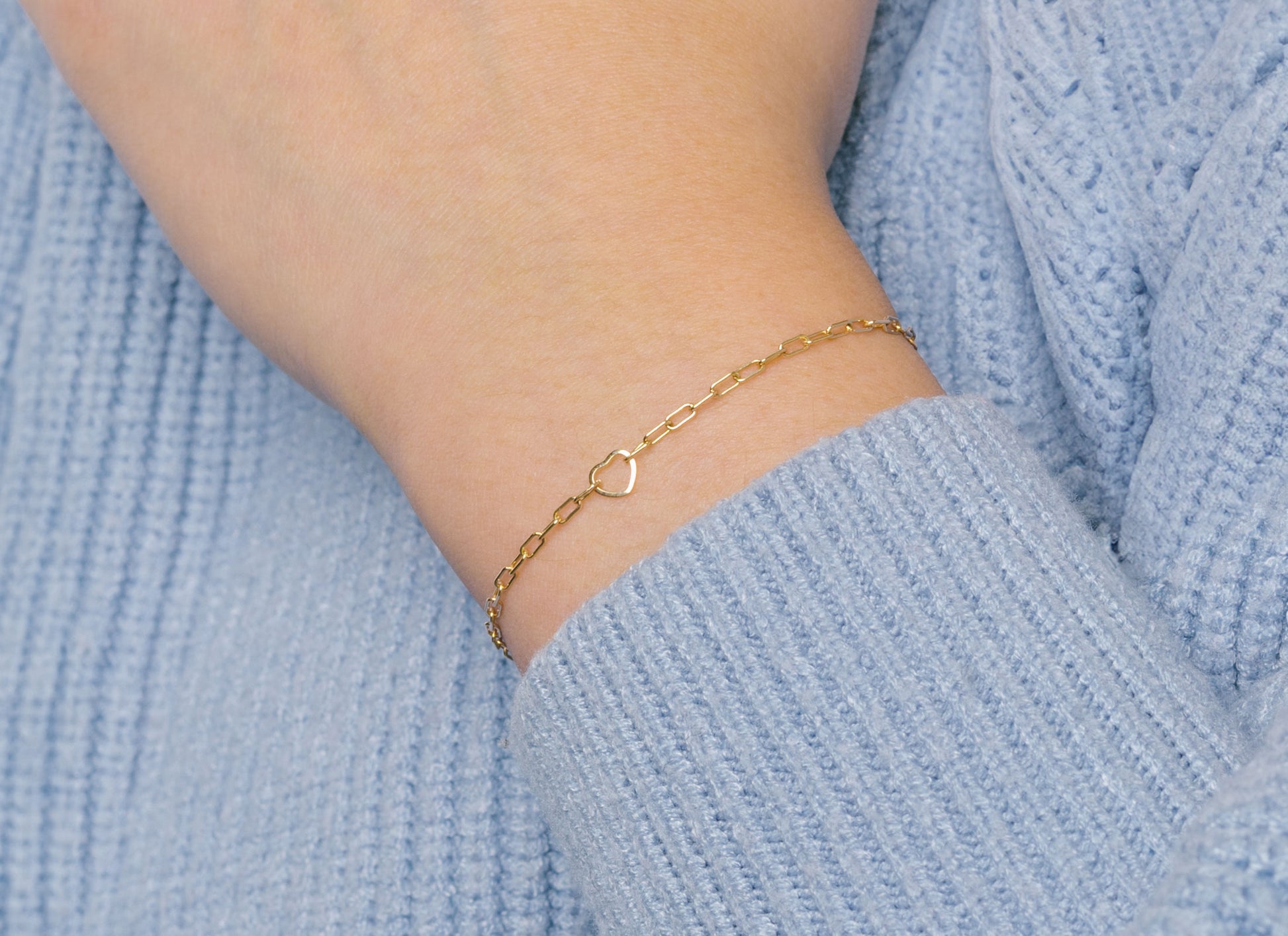 A gold heart-shaped wire charm connector worn as a bracelet on a person's wrist, with a sweater in the background.