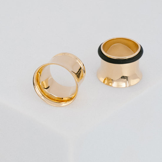 Two gold ear gauges with black rubber tips on a light gray background