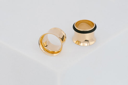 Two gold ear gauges with black rubber tips on a light gray background