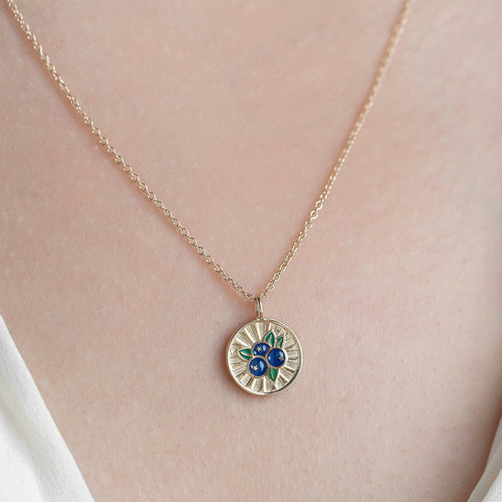 Gold necklace with a circular pendant featuring blueberries on a person wearing a white top.